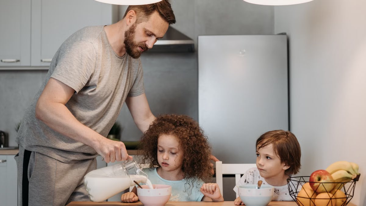 A father pours milk into cereal bowls for his children at the breakfast table in a modern kitchen.